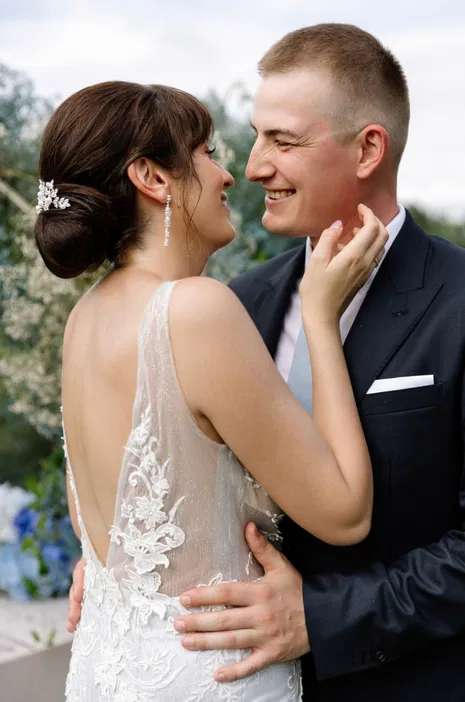 Para młoda obejmująca się podczas wesela, uśmiechnięci, ona w koronkowej sukni ślubnej, on w eleganckim garniturze, romantyczna scena na świeżym powietrzu.