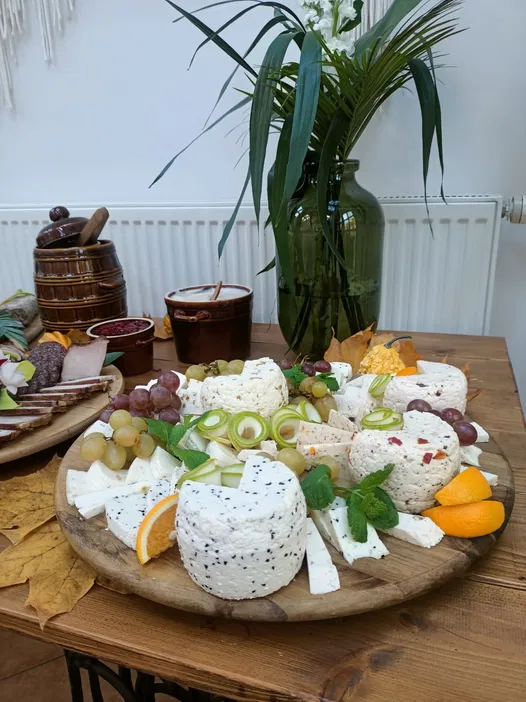 Serowa deska z różnymi rodzajami sera, plasterkami pomarańczy, winogronami i innymi ozdobami na drewnianym stole, z wazą i rośliną w tle.