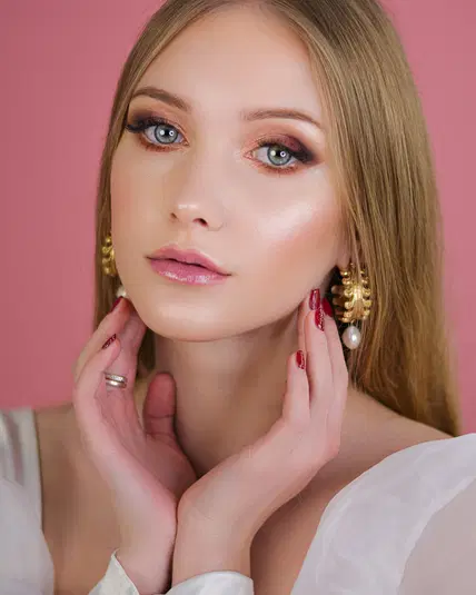  Blondynka z subtelnym makijażem i złotymi kolczykami na różowym tle, dotykająca twarzy dłońmi z czerwonym lakierem na paznokciach. 