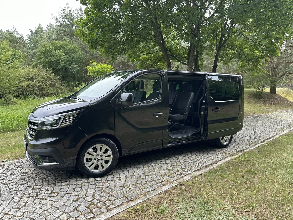 Czarny van z otwartymi drzwiami na tle zielonego parku, auto stoi na brukowanej drodze.