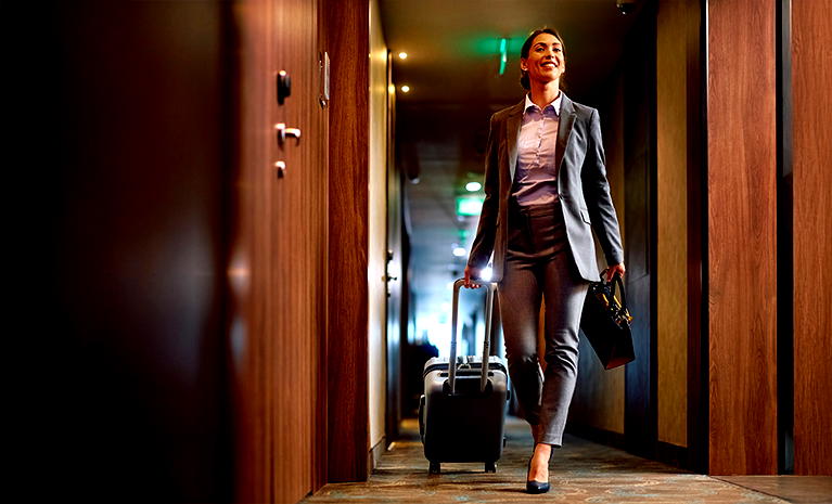  Kobieta w eleganckim garniturze z walizką na kółkach, idąca korytarzem hotelowym. 