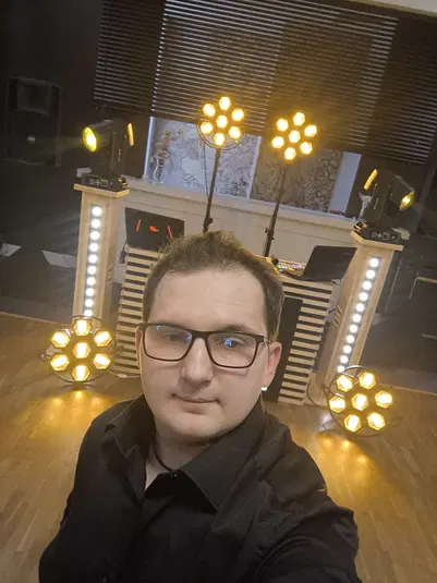  DJ w czarnej koszuli na tle profesjonalnego sprzętu oświetleniowego w sali eventowej. 