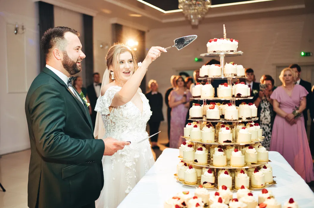 Para młoda krojąca tort weselny, uroczystość ślubna, elegancki tort na stojaku, radosna atmosfera, goście weselni w tle.
