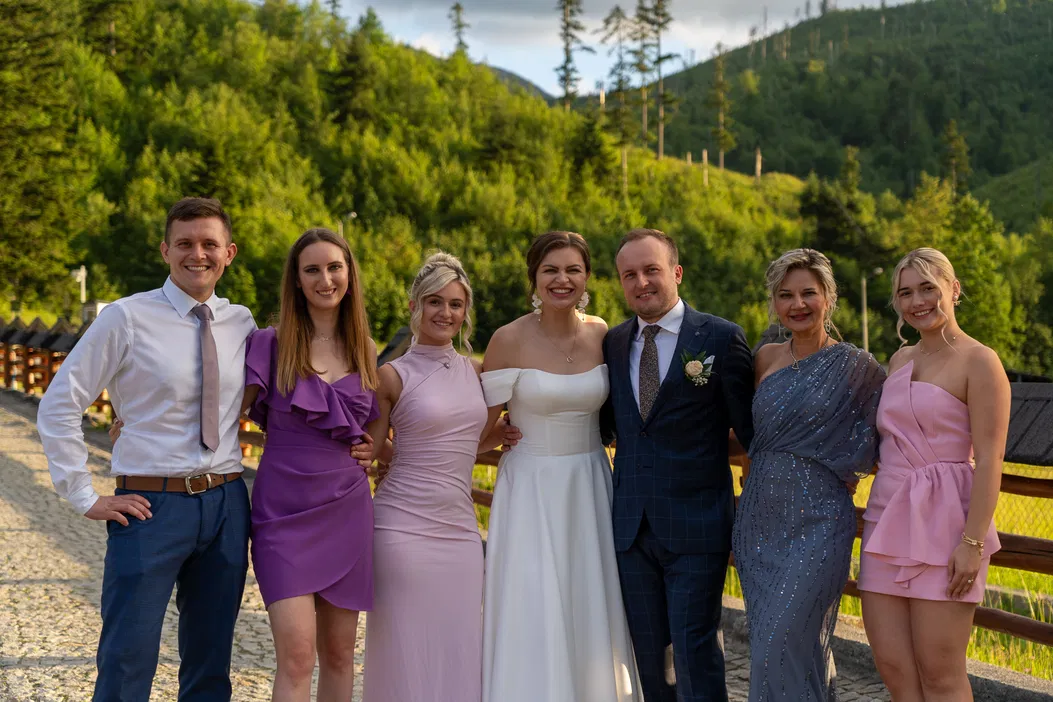 Grupa uśmiechniętych ludzi ubranych w eleganckie stroje stoi na tle zielonych gór i lasu, uczestniczą w przyjęciu na świeżym powietrzu.