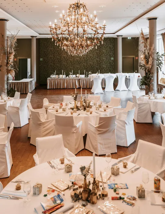 Elegancka sala weselna z luksusowym żyrandolem, białymi obrusami na okrągłych stołach i dekoracjami ślubnymi.