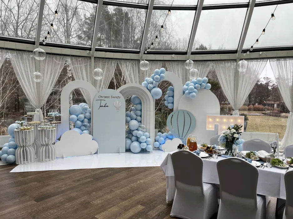 Dekoracje na chrzest święty z niebieskimi balonami i stołem pełnym jedzenia w eleganckim wnętrzu z przeszklonym dachem.