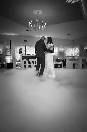  Para tańczy pierwszy taniec na weselu, otoczona mgłą, eleganckie wnętrze z żyrandolem, napis LOVE w tle. 