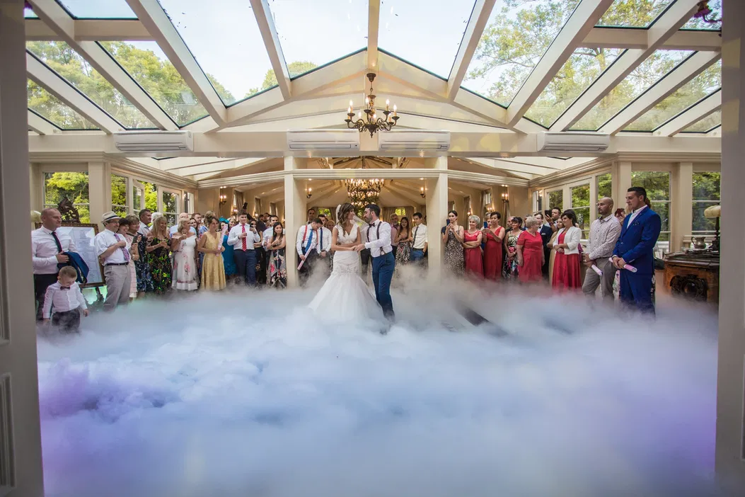 Pierwszy taniec pary młodej w otoczeniu gości weselnych w eleganckiej, przeszklonej sali z efektem mgły na podłodze.