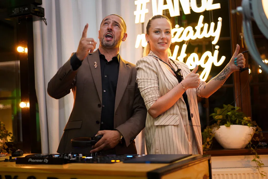 DJ prowadzący imprezę w eleganckim garniturze obok kobiety w jasnej marynarce, z neonowym napisem w tle.