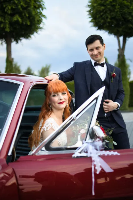 Alternatywny tekst: Para młoda przy klasycznym samochodzie, panna młoda w koronkowej sukni, pan młody w eleganckim garniturze, wesele na świeżym powietrzu.