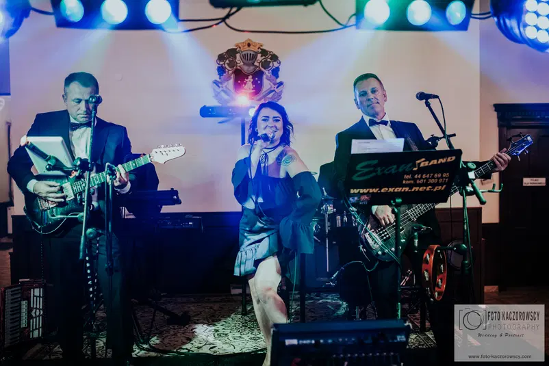  Zespół muzyczny grający na żywo podczas występu na scenie, trzech członków zespołu w eleganckich strojach, gitara, mikrofon, kolorowe oświetlenie. 