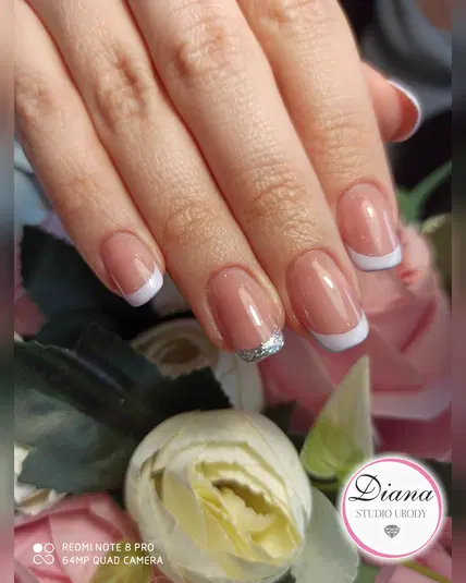  Elegancki manicure francuski z białymi końcówkami i srebrnym akcentem, wykonany w Studio Urody Diana na naturalnych paznokciach, na tle kwiatów. 