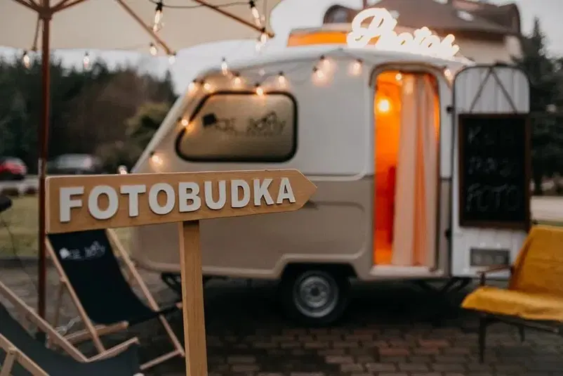  Przyczepa kempingowa z fotobudką, oznaczona drewnianym szyldem, otoczona dekoracyjnymi lampkami, przedmieścia, wieczorny klimat. 