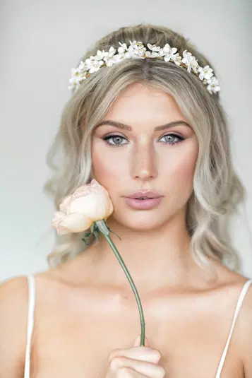  Blondynka z wieńcem kwiatowym na głowie trzymająca różę, delikatny makijaż, romantyczny styl ślubny. 