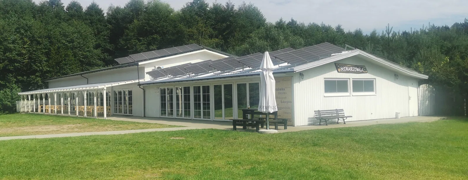 Budynek z panelami słonecznymi otoczony zielenią, widok z zewnątrz, altana z drewnianymi ławkami, słoneczny dzień, ekologiczna architektura, nowoczesne rozwiązania energetyczne.