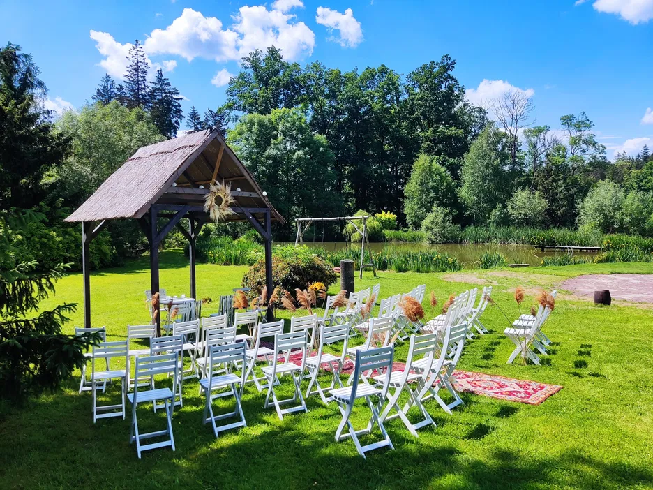 Plenerowa ceremonia ślubna w zielonym ogrodzie z białymi krzesłami, drewnianą altaną i malowniczym krajobrazem