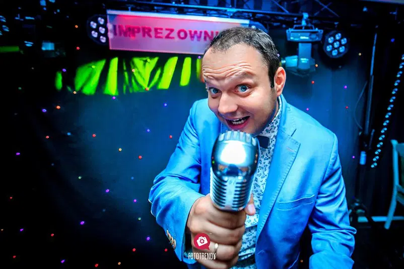  Mężczyzna w niebieskiej marynarce trzymający mikrofon, scena z kolorowymi światłami w tle, napis IMPREZOWNIA╗ 