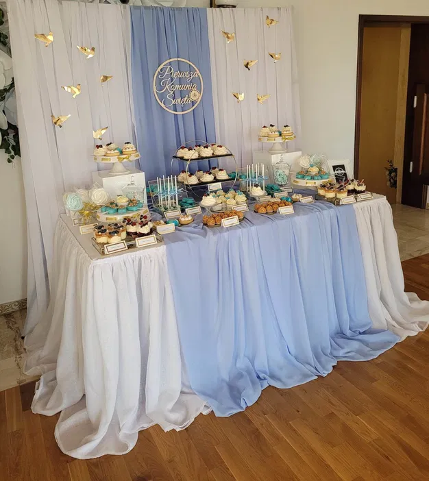 Dekoracja stołu na Pierwszą Komunię Świętą z niebieskimi i białymi słodyczami, babeczkami i kwiatami na tle zasłon w pastelowych kolorach oraz napisem Pierwsza Komunia Święta.