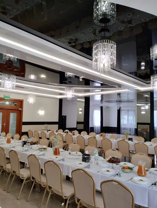 Elegancka sala bankietowa z ustawionymi stołami, przygotowana na przyjęcie; dekoracyjne żyrandole, białe obrusy, zastawa, krzesła przy stołach.