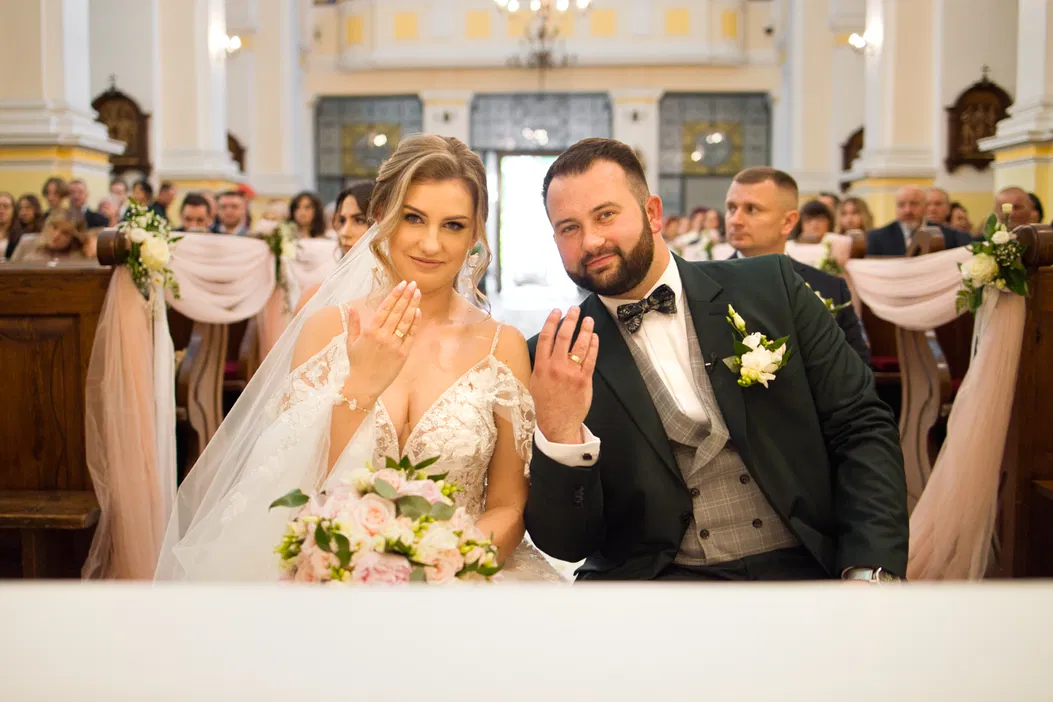 Para młoda w kościele podczas ceremonii ślubnej, prezentująca obrączki ślubne; panna młoda w koronkowej sukni z welonem trzyma bukiet kwiatów; elegancki pan młody w garniturze.