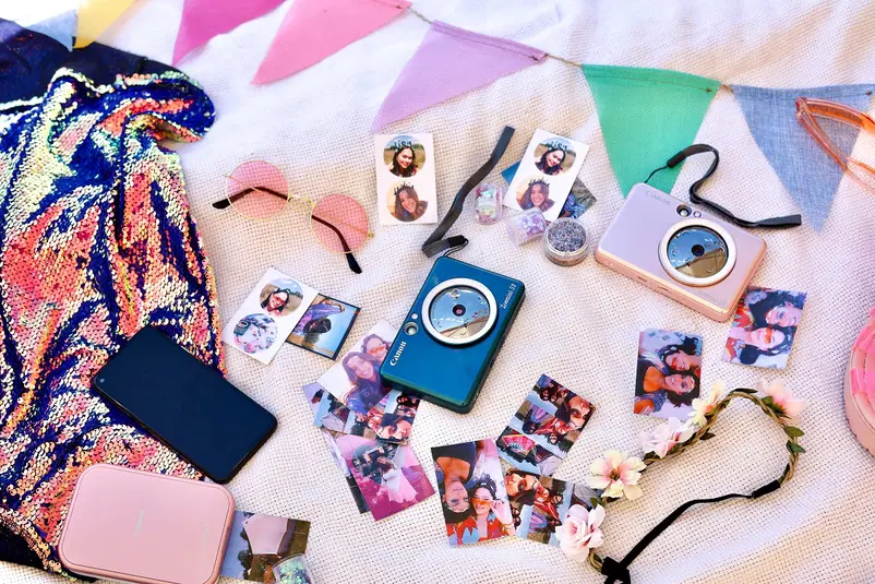 Festynowe akcesoria na piknikowym kocu: polaroidy, błyszczące aparaty, okulary przeciwsłoneczne, girlanda, cekinowa tkanina. 