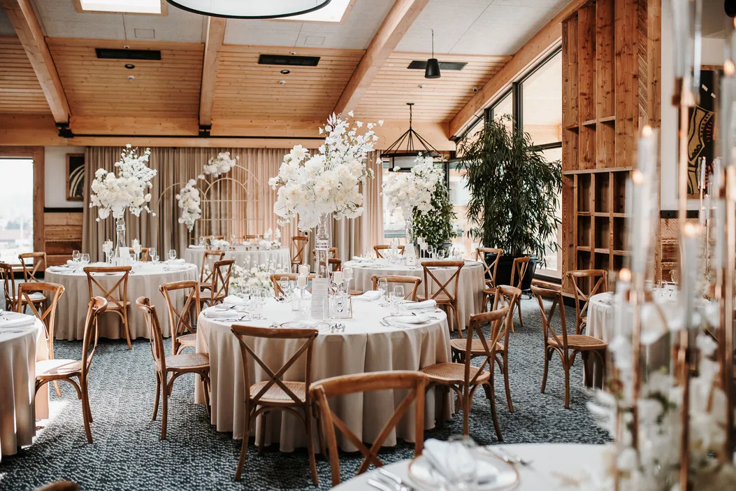 Elegancka sala weselna z okrągłymi stołami, białymi obrusami i kwiatowymi dekoracjami. Drewniane krzesła, nowoczesne oświetlenie i duże okna tworzą przytulną atmosferę. Idealne miejsce na wesele.
