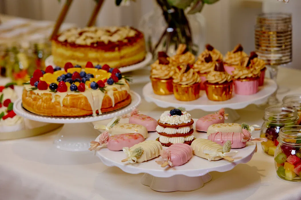 Stół z deserami: tort z owocami, babeczki w złotych papilotkach, eleganckie ciasteczka z polewą i deserki w słoikach.