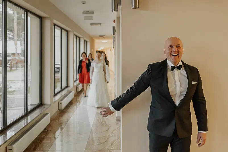  Mężczyzna w garniturze z uśmiechem na korytarzu, w tle kobiety w eleganckich strojach z panną młodą w białej sukni, emocjonalna chwila na weselu. 