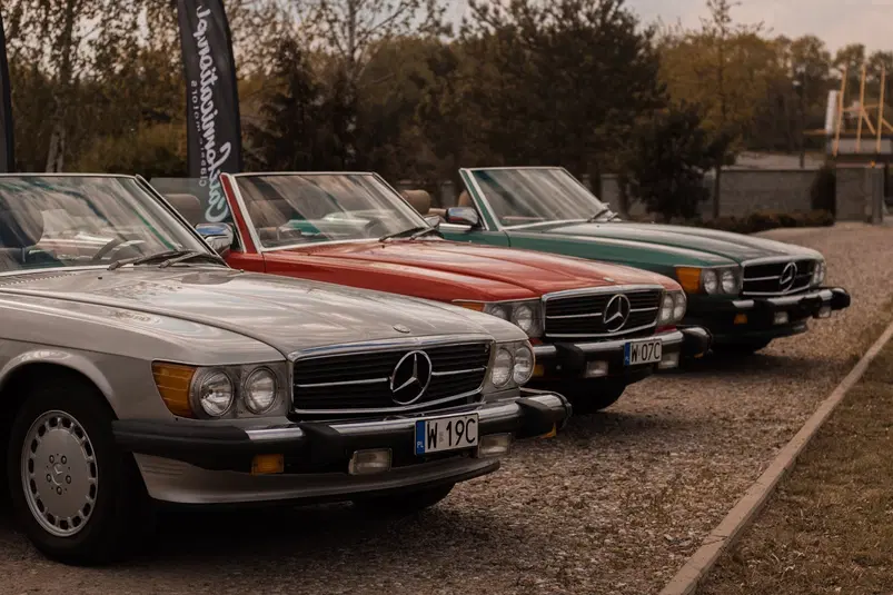  Trzy klasyczne samochody marki Mercedes-Benz w różnych kolorach zaparkowane na żwirowym podjeździe, otoczone drzewami. 