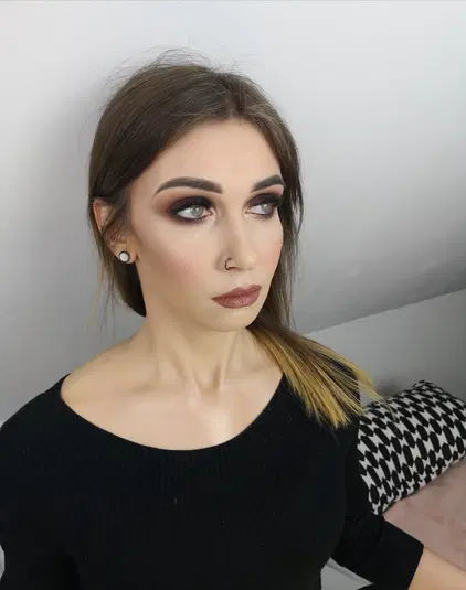  Ujęcie kobiety z ciemnym makijażem oczu i nude ustami, ubranej w czarny sweter, na jasnym tle. 