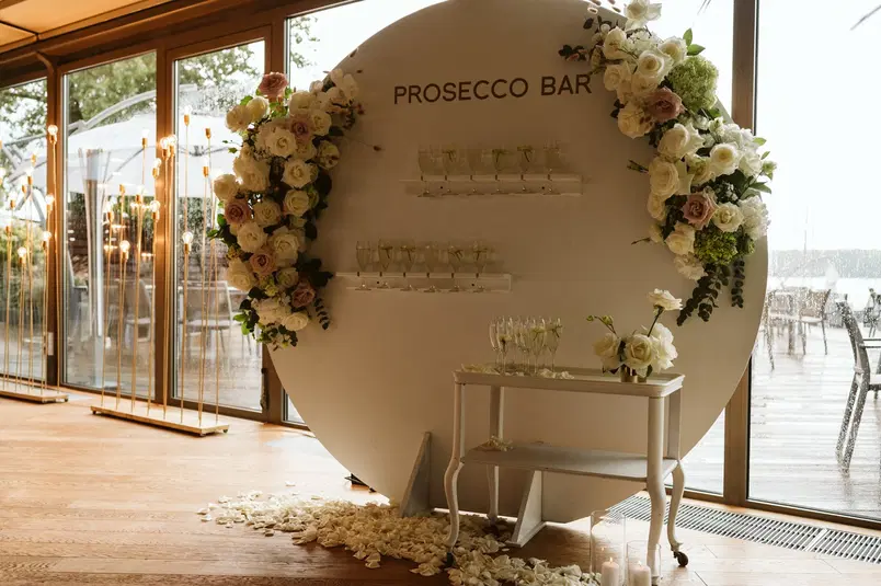  Alta zdjęcia: Stylowy bar prosecco z dekoracją kwiatową na eleganckim przyjęciu we wnętrzu z drewnianą podłogą i przeszklonymi drzwiami, serwujący kieliszki musującego wina. 