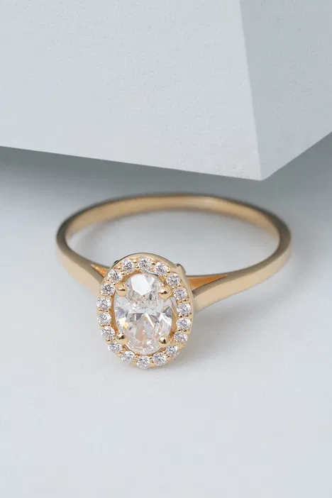 Złoty pierścionek z owalnym diamentem otoczonym mniejszymi diamentami, elegancka biżuteria na białym tle.
