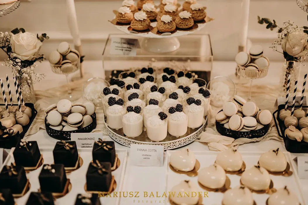 Stół z eleganckimi deserami, w tym panna cotta z jeżynami, makaroniki karmelowe, tarta czekoladowa i ozdobne ciasta, idealne na przyjęcia i wesela.