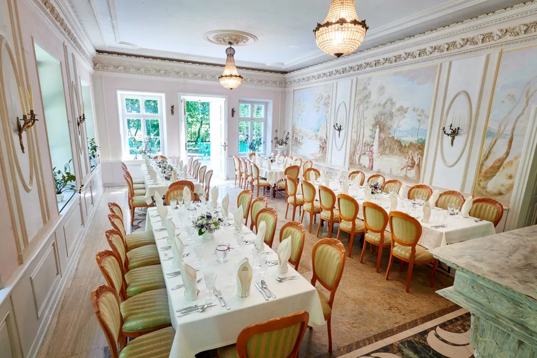Luksusowa sala bankietowa z elegancko nakrytymi stołami, klasyczne żyrandole, dekoracyjne freski na ścianach, krzesła w stylu retro, jasne wnętrze z dużymi oknami, idealna na przyjęcia i uroczystości.