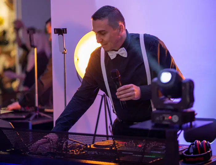  DJ na imprezie z mikrofonem, ubrany elegancko, przy sprzęcie muzycznym, intensywne oświetlenie sceny, profesjonalna obsługa wydarzeń muzycznych. 