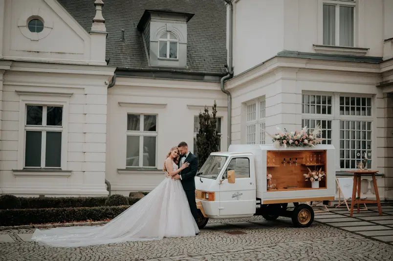  Para młoda przed eleganckim budynkiem w pobliżu mobilnego baru z prosecco, ozdobionego kwiatami - luksusowe wesele na świeżym powietrzu. 