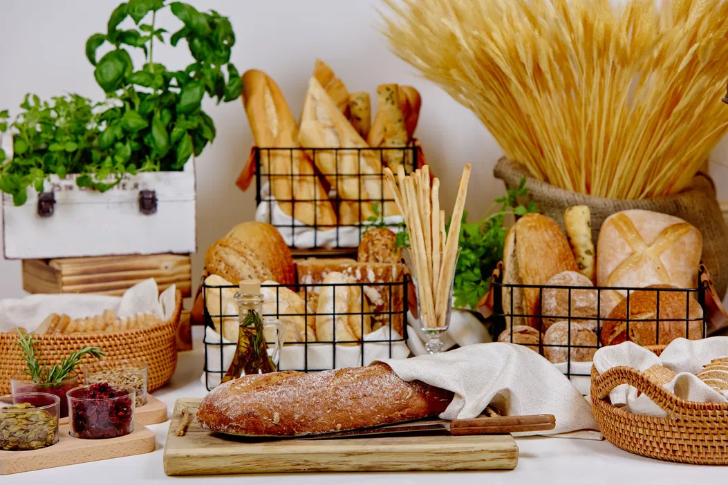 Stół z różnymi rodzajami pieczywa, bagietkami, chlebem razowym i ziołami, dekoracja z pszenicą w tle, rustykalny wystrój kuchni.
