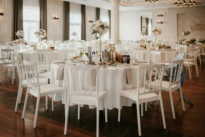  Elegantna sala weselna z okrągłymi stołami, białymi krzesłami i kwiatowymi dekoracjami, przygotowana na przyjęcie weselne. 