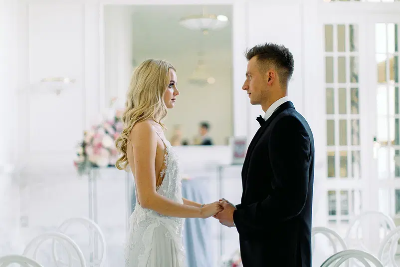  Para w strojach ślubnych trzymająca się za ręce podczas ceremonii w eleganckiej, jasnej sali. 