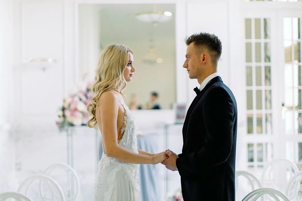 Para w strojach ślubnych trzymająca się za ręce podczas ceremonii w eleganckiej, jasnej sali.