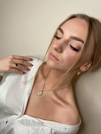  Młoda kobieta w białej bluzce z subtelnym makijażem i biżuterią, złoty naszyjnik i kolczyki, elegancka stylizacja. 