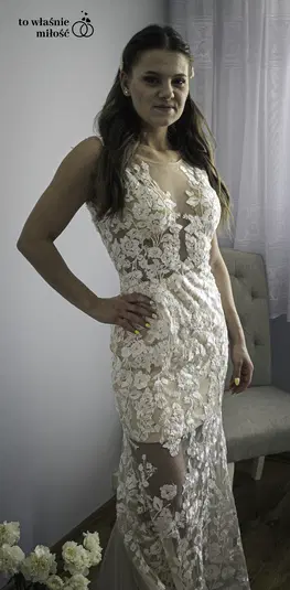  Kobieta w koronkowej sukni ślubnej w eleganckim wnętrzu, kwiaty i zasłony w tle. 