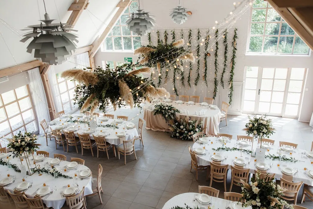 Elegancka sala weselna z dekoracjami kwiatowymi i białymi obrusami, przygotowana na przyjęcie weselne, duże okna wpuszczają naturalne światło do wnętrza.