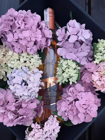  Butelka wina HR Winery Chardonnay otoczona bukietem różowych i jasnofioletowych hortensji, elegancka kompozycja w czarnym pudełku 