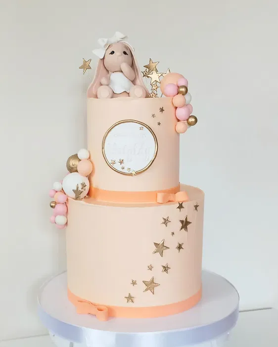Dwupiętrowy tort w pastelowych kolorach z dekoracjami w kształcie gwiazd i figurką króliczka na górze, idealny na uroczystość chrztu.
