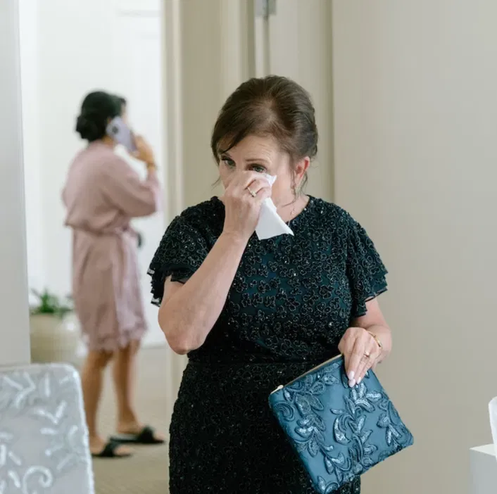 Kobieta w eleganckiej sukience wyciera łzy podczas przygotowań do ważnego wydarzenia, w tle inna osoba rozmawiająca przez telefon.