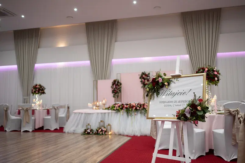  Wystawne dekoracje weselne, elegancka sala bankietowa, kwiatowe aranżacje, ozdobny napis powitalny na stojaku, białe pokrowce na krzesła, romantyczne oświetlenie. 