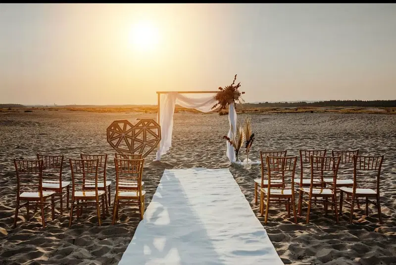  Plażowa ceremonia ślubna o zachodzie słońca, udekorowana elegancko krzesłami, łukiem i suszonymi kwiatami. 