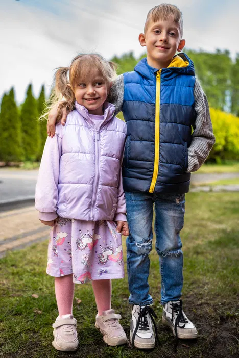 Dwoje dzieci ubranych w zimowe kurtki, stojące na trawie na tle drzew, chłopiec w niebieskiej puchowej kamizelce i dziewczynka w różowej kurtce.