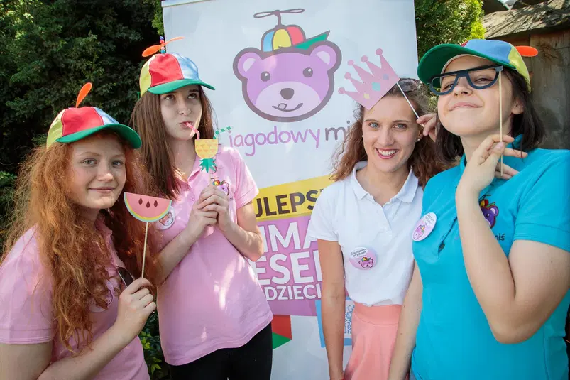  Cztery młode osoby w kolorowych czapkach z płaskim śmigłem pozują przed banerem Jagodowy Miś podczas letniej imprezy plenerowej. 